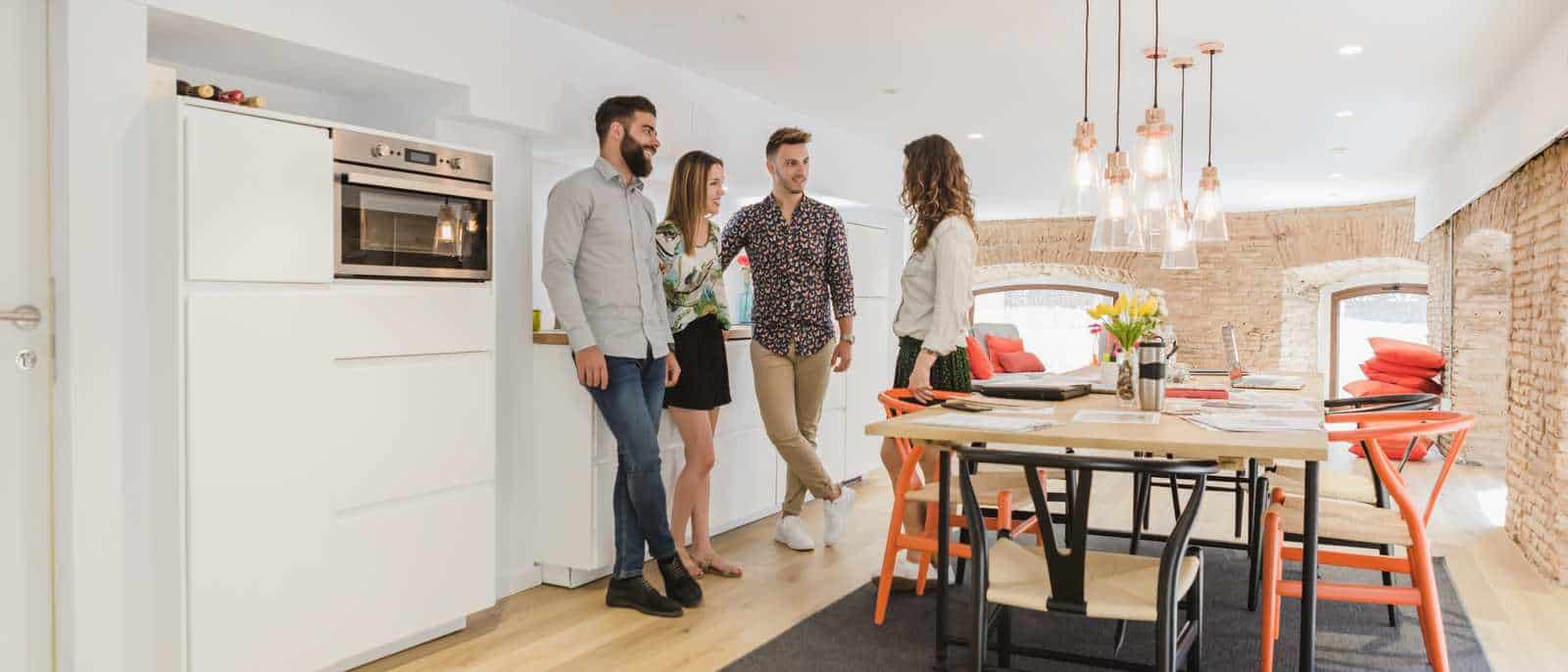 Quatre jeunes adultes debout dans une cuisine ouverte sur une salle à manger, mur de brique et suspensions au-dessus de la table.