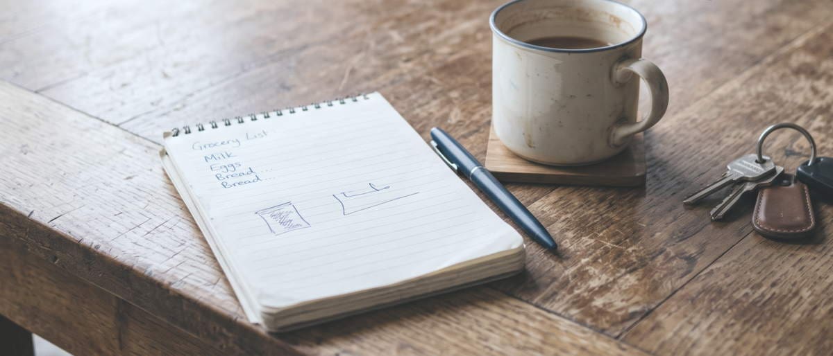 Un carnet ouvert avec une liste écrite à la main, un stylo et une tasse de café sur une table en bois usée.