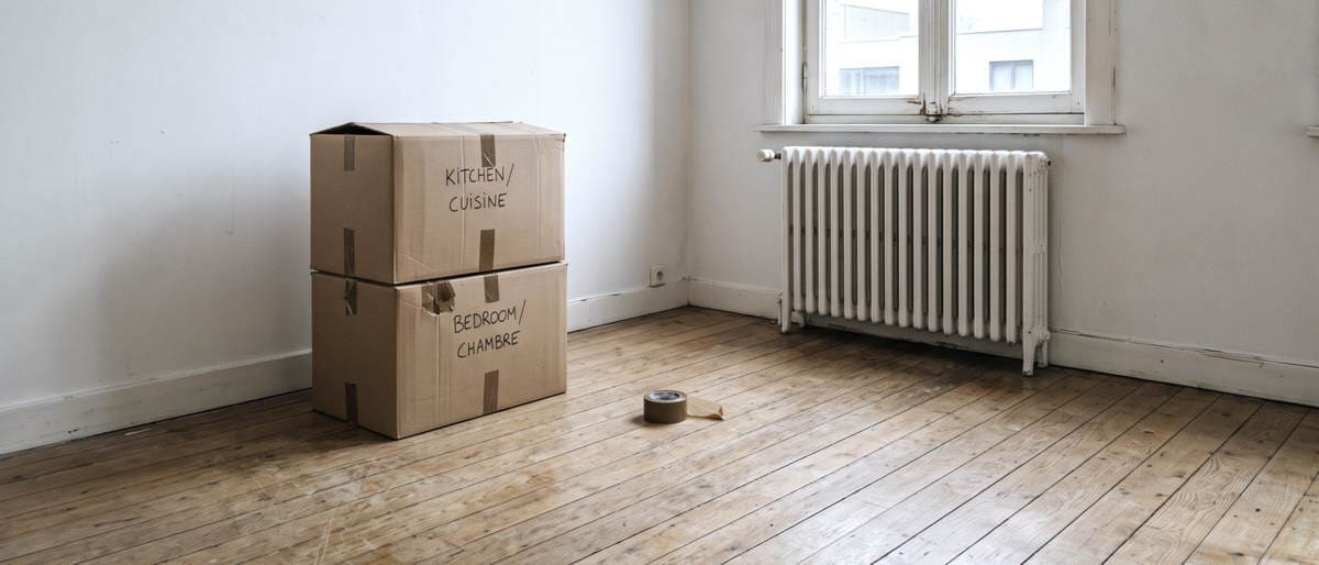 Deux cartons de déménagement empilés contre un mur blanc dans une pièce vide, un rouleau d'adhésif posé sur le parquet, un radiateur sous la fenêtre.