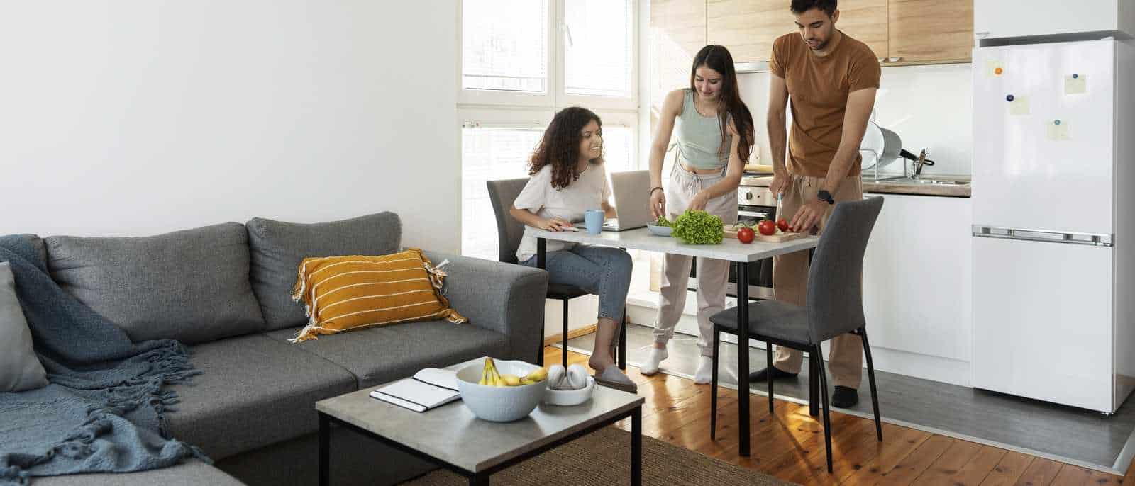 Trois jeunes adultes dans le séjour d'un appartement partagé, autour d'une table près du coin cuisine, canapé et table basse au premier plan.