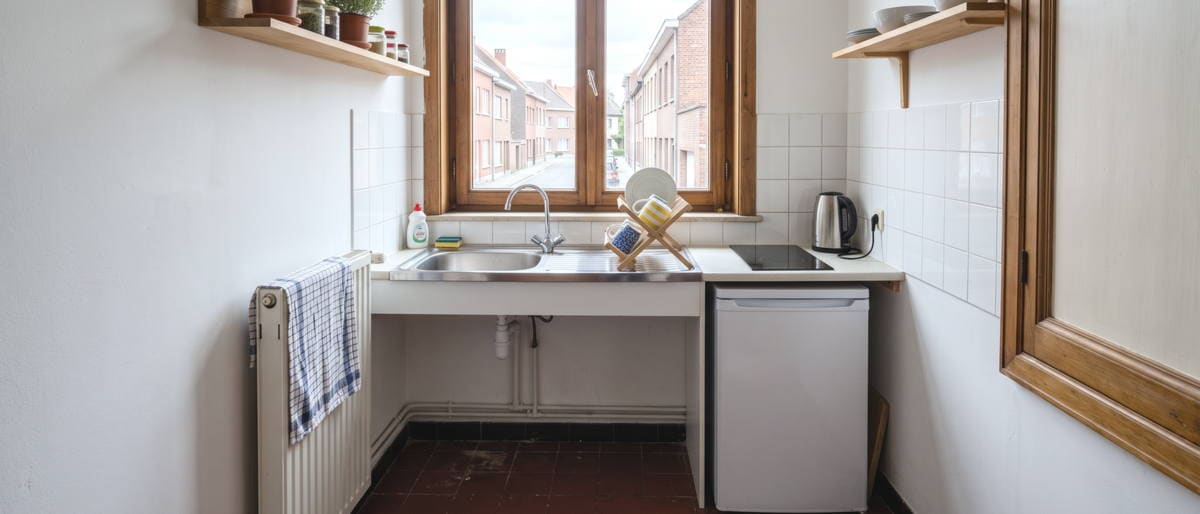 Une petite cuisine commune de kot, plan de travail étroit avec une bouilloire et un égouttoir, un petit frigo et une fenêtre au-dessus de l'évier.