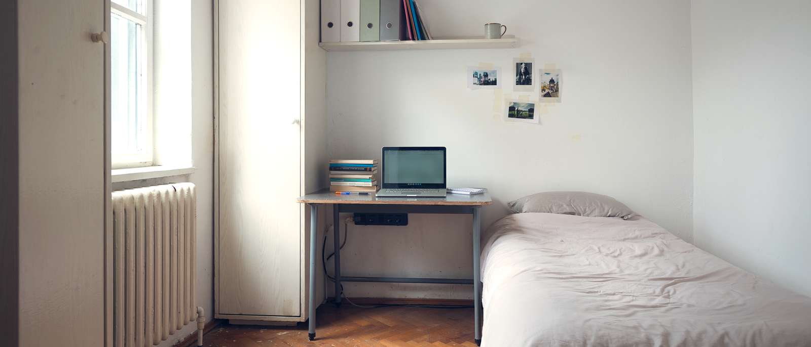 Une chambre d'étudiant, lit simple, bureau avec un ordinateur portable et une pile de livres, radiateur sous la fenêtre et parquet ancien.