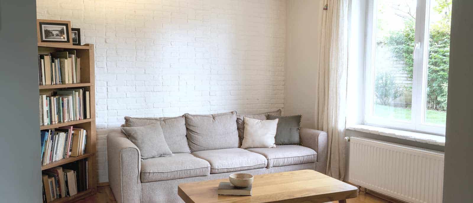 Le salon d'une maison en location, mur de brique peint en blanc, canapé clair, bibliothèque et table basse devant une fenêtre ouverte sur un jardin.