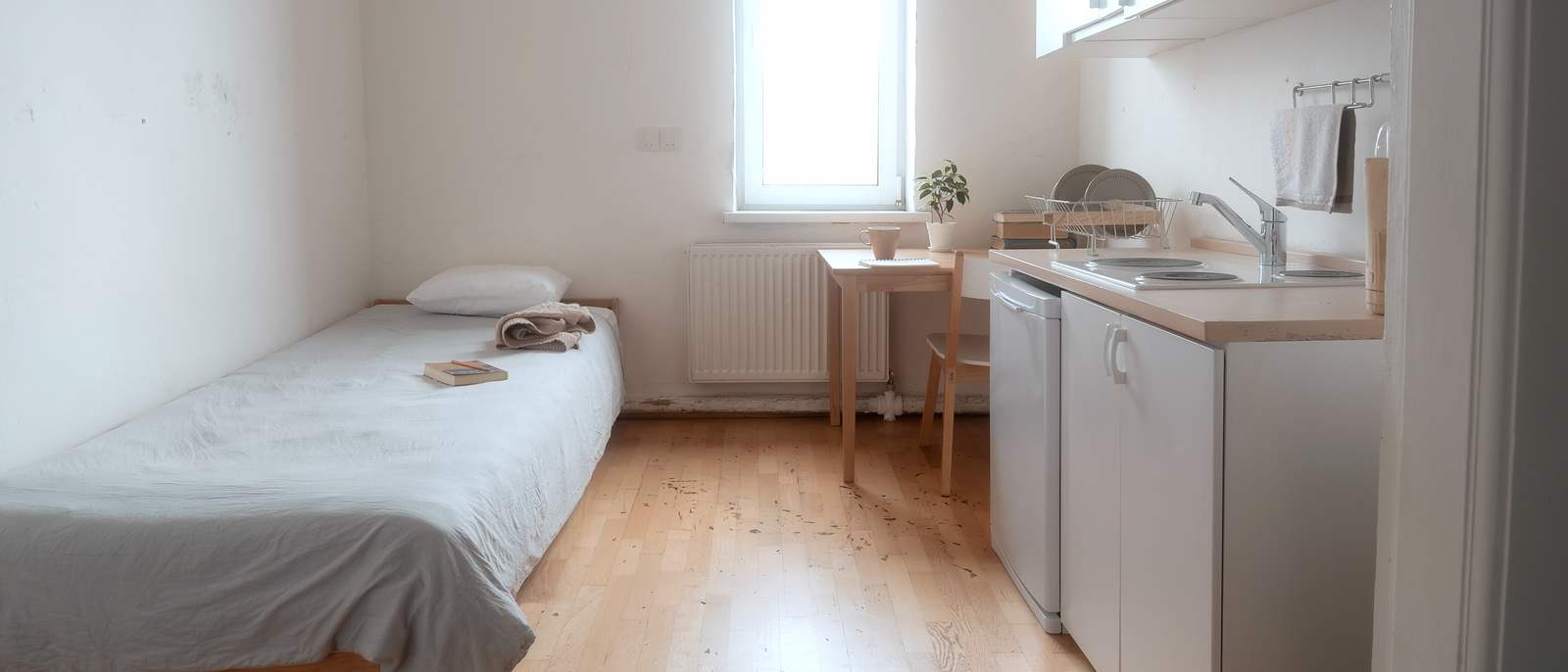 Un studio d'une seule pièce, lit simple contre le mur, petite cuisine avec évier et frigo, table sous la fenêtre et parquet clair.